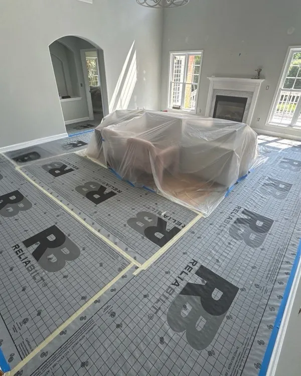 A living room with the floor covered in synthetic underlayment, edges taped and the sofa wrapped in plastic